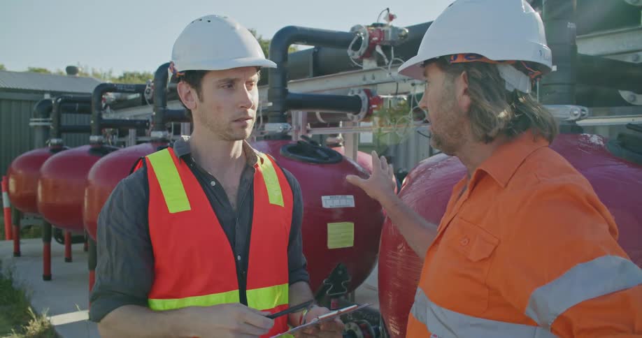 Two men standing outdoors at daytime wearing hard hats and safety vest having a conversation while one takes notes on a clipboard