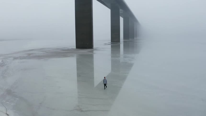 Man walking over the frozen surface of a lake under a bridge on a foggy day