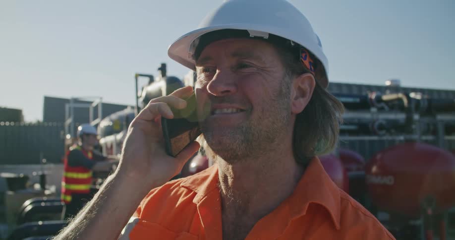 A man with a hardhat talking on a smart phone with the sky and clouds in the background