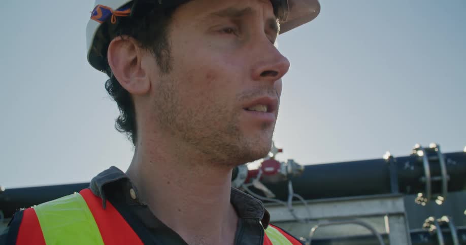 A man having a conversation with his coworker outdoors at a manufacturing plant while a flare of sunlight shines from behind him