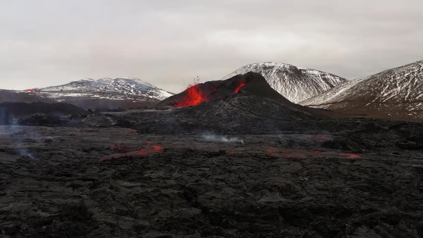 Erupting volcano spews molten hot lava into the air with snow covered mountains in the distance