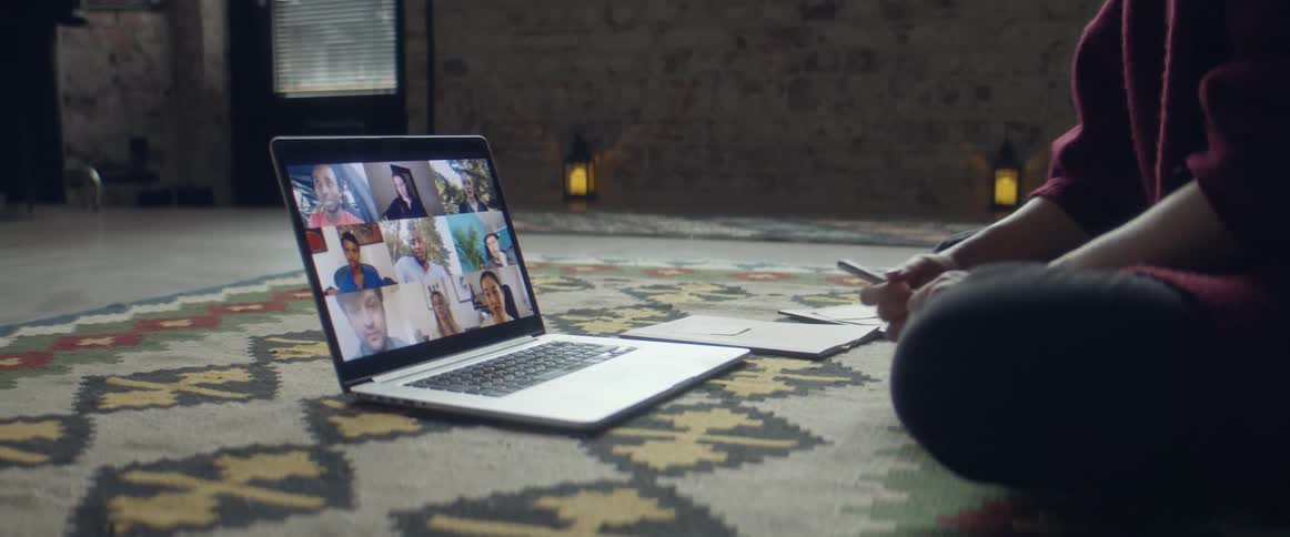 A person sits in front of a laptop on the floor having a video call and writing in a notebook