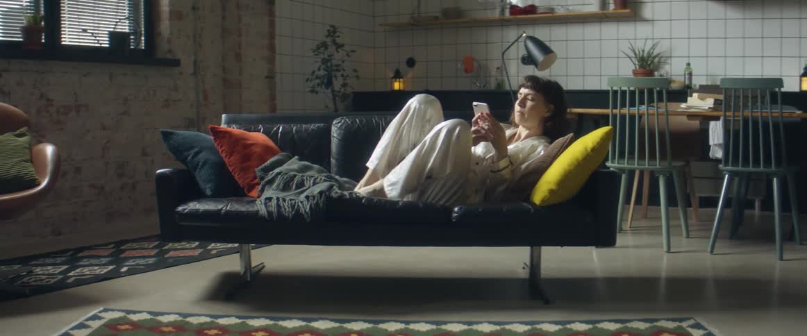 Woman wearing pajamas reclines on sofa with smartphone near kitchen table and window