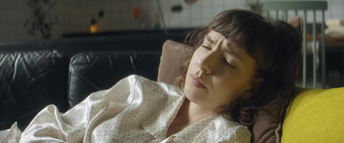 A woman scrolls through her phone while lying on a couch indoors then puts the phone down and stares off with a worried expression