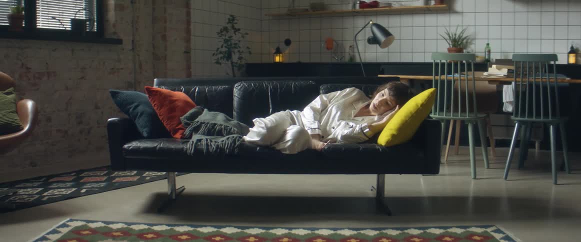 A woman checks her smartphone before lying back to rest on a sofa in her pajamas at home during the day
