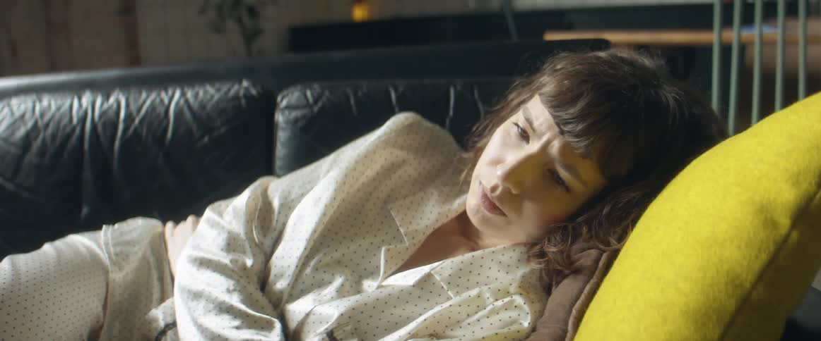A woman in pajamas laying on a couch with a wall and a chair in the background