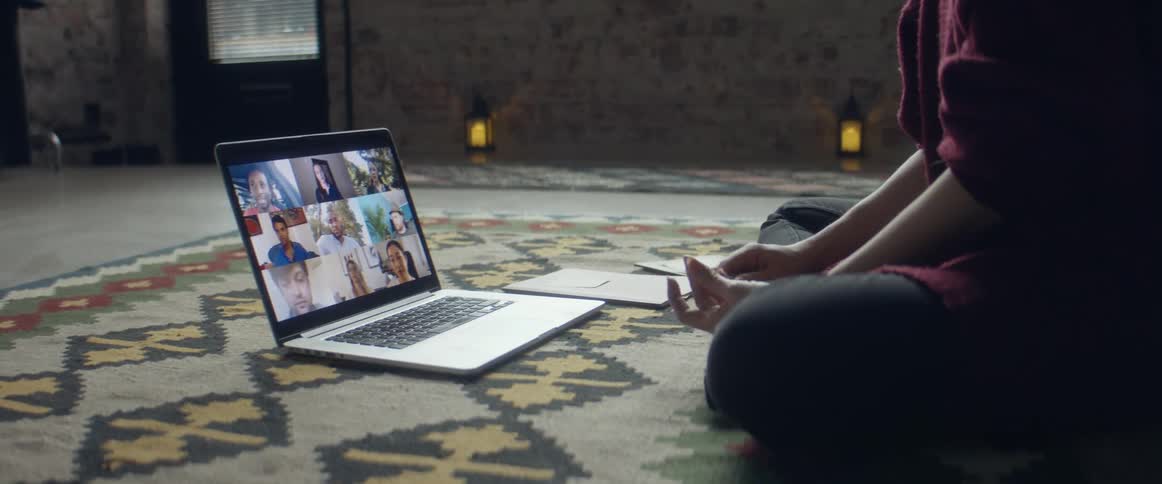 A teenager sits on the floor at home and video chats on a laptop