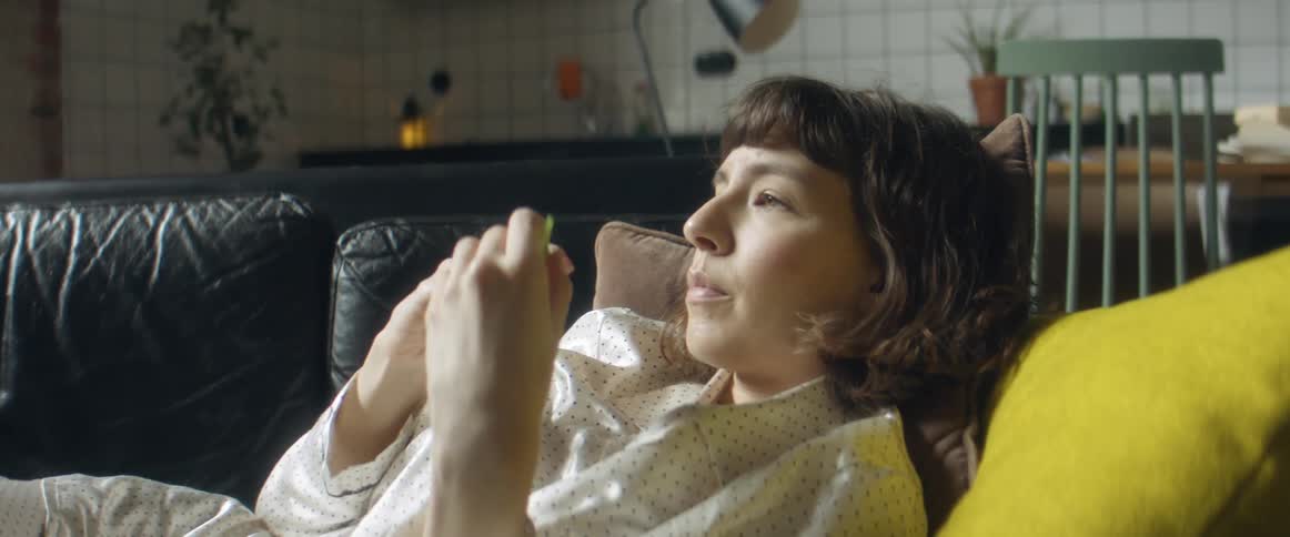 A woman laying down on a couch at home during the day puts her cell phone down and takes a thermometer from under her arm and checks her temperature