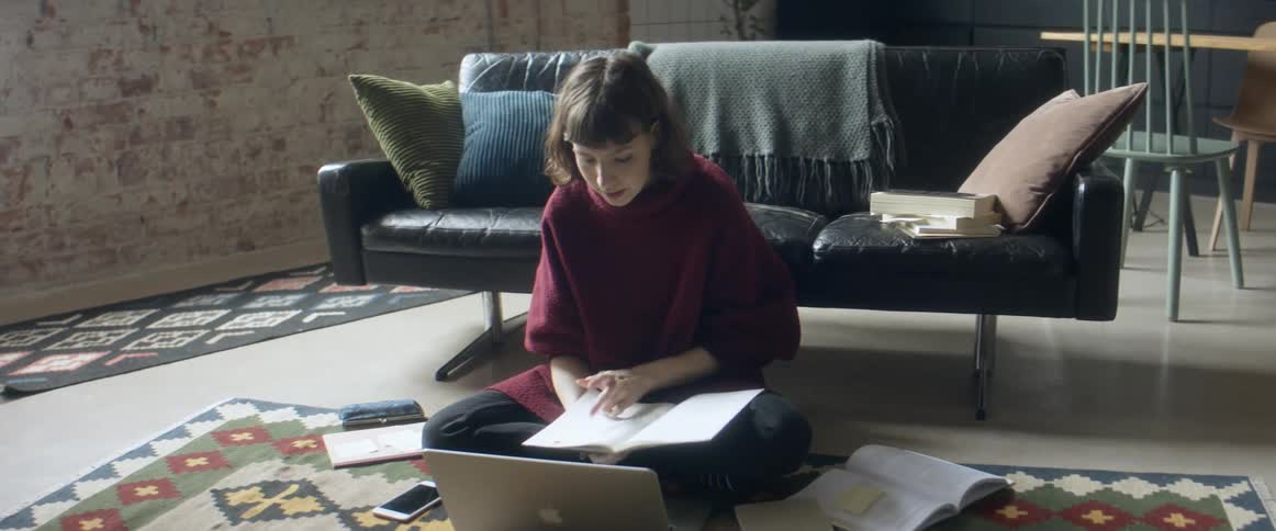 A woman sits on the floor while looking through a notebook as she talks to someone via her laptop
