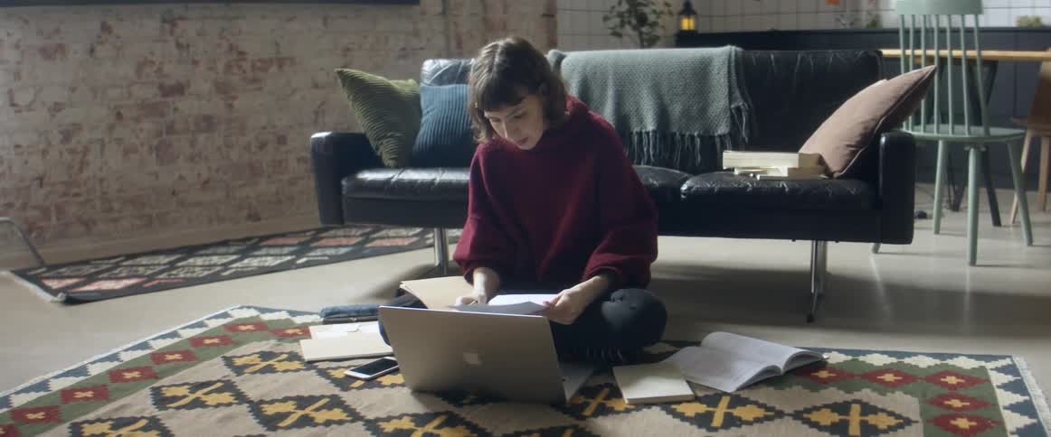 A woman talks to someone on her laptop while studying and sitting on the floor of her living room
