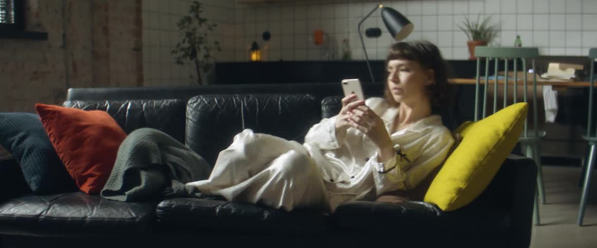 A woman walks across a room to rest on a sofa and check her smartphone