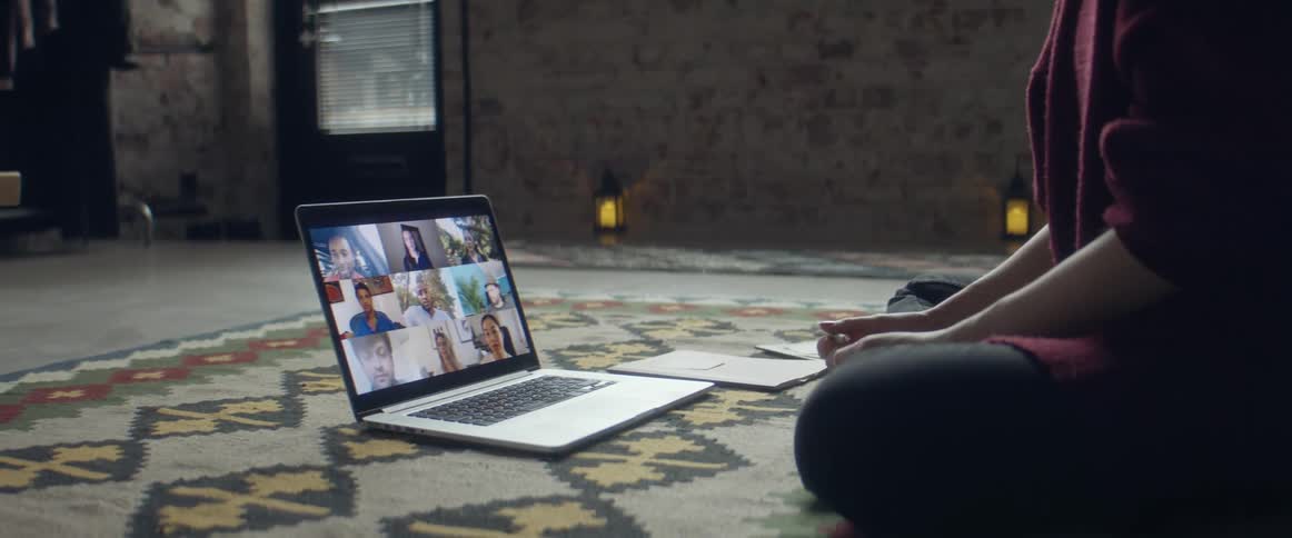 A woman sitting on a rug in a living room taking notes in a notebook while on a zoom call on a laptop with other people
