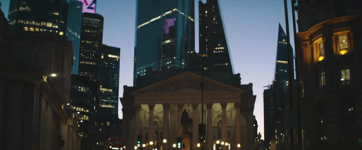 A double decker bus is stopped downtown below tall skyscrapers lit up at night