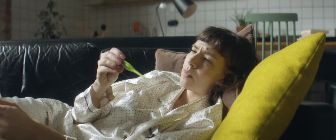 A woman lying on a couch puts her phone down and takes the thermometer out from under her arm