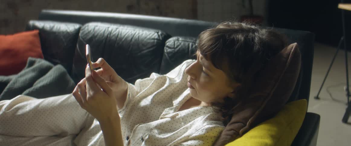 A white female in pajamas is on a couch laying down using a cell phone