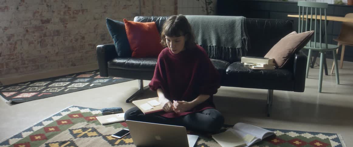 A woman sitting on the floor on a carpet in front of a laptop with a couch and wall in the background