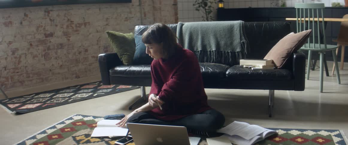 A woman sits on the floor in front of a laptop and also looking through a book