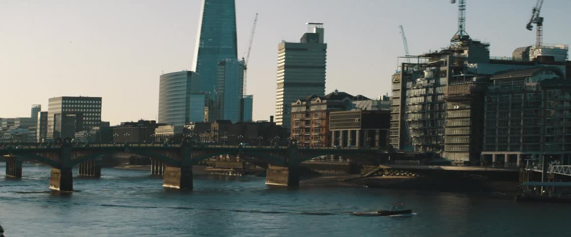 A boat in a river as cars drive across a bridge with buildings and construction cranes in the background