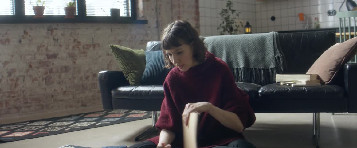 A woman sitting on the floor talks and nods at her laptop while flipping through a notebook