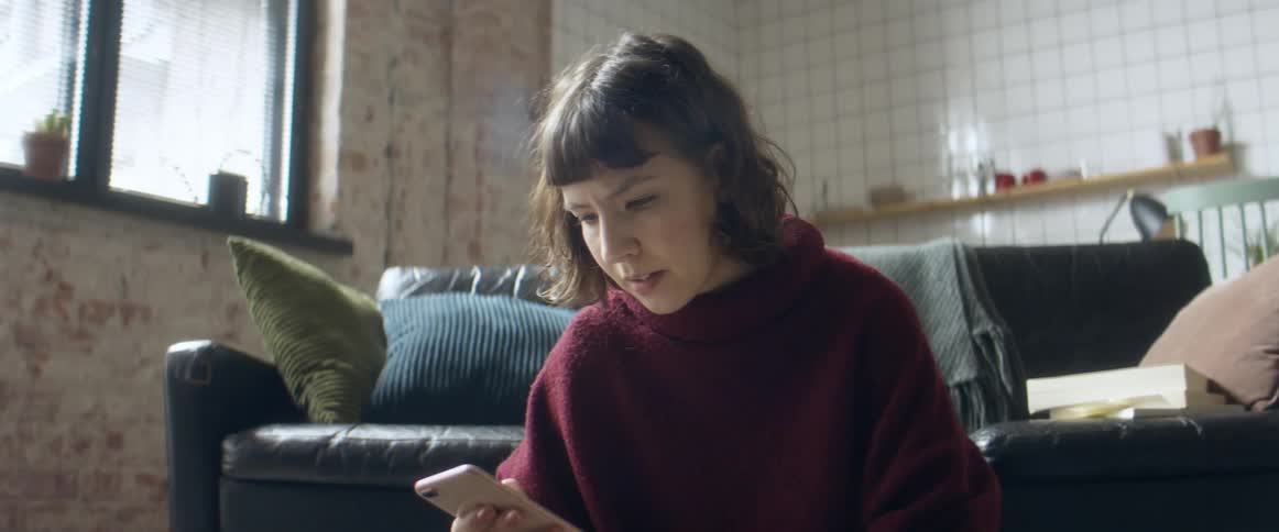 A woman sits on the floor of her living room with her laptop while using her smartphone