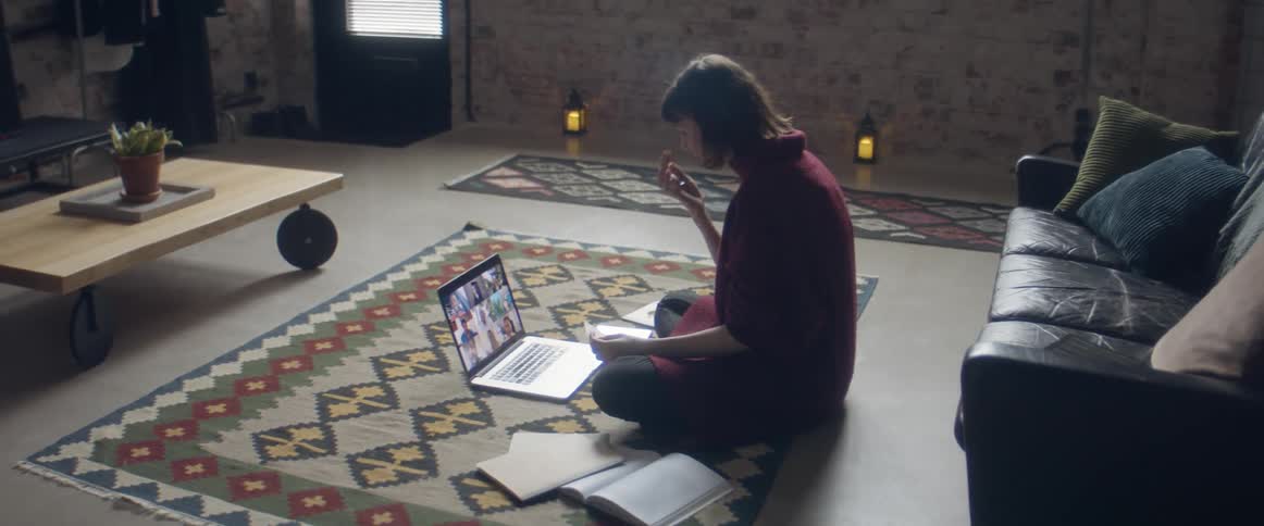 A woman sits on the floor with an open notebook as she participates in a video conference via her laptop
