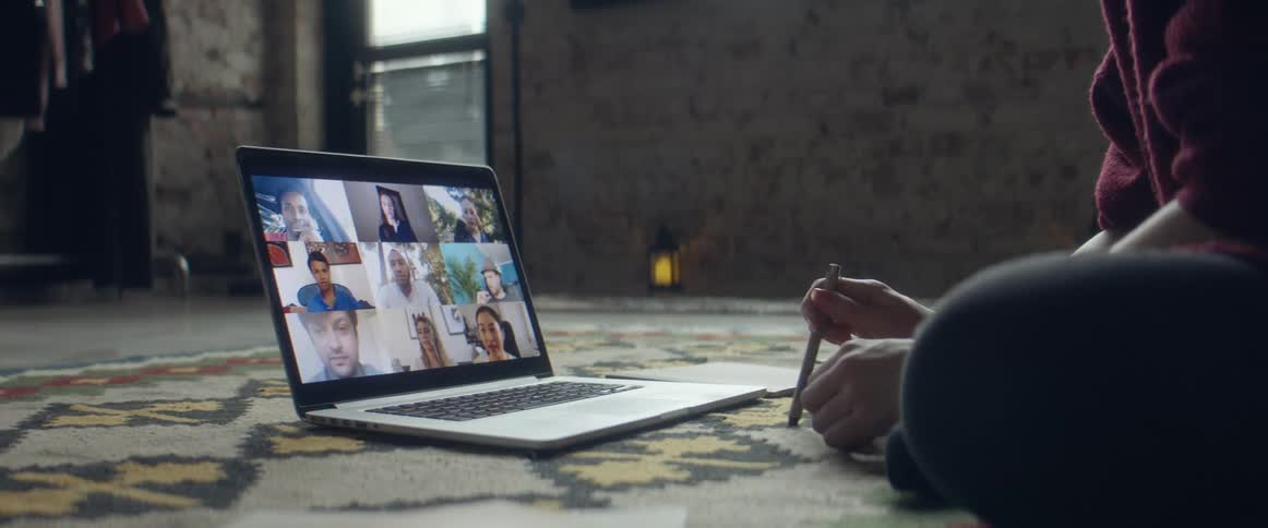 A woman talking to a group of friends in a zoom call on a laptop computer on the floor in a living room