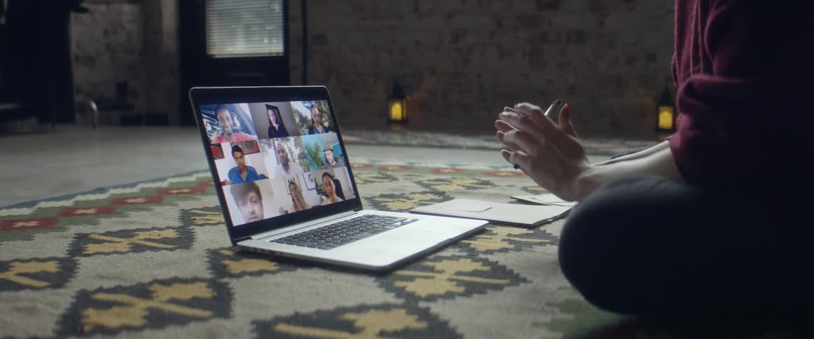 A woman sitting on the floor gestures as she talks during a video call on her laptop