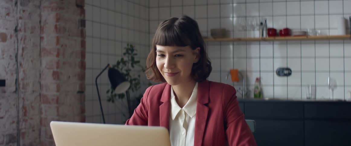 A woman in a room sitting on a chair in front of a laptop with a wall in the background