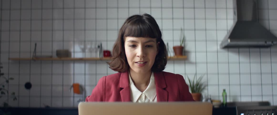 Woman wearing business jacket teleconferences via laptop sitting at home with white tiled kitchen as backdrop