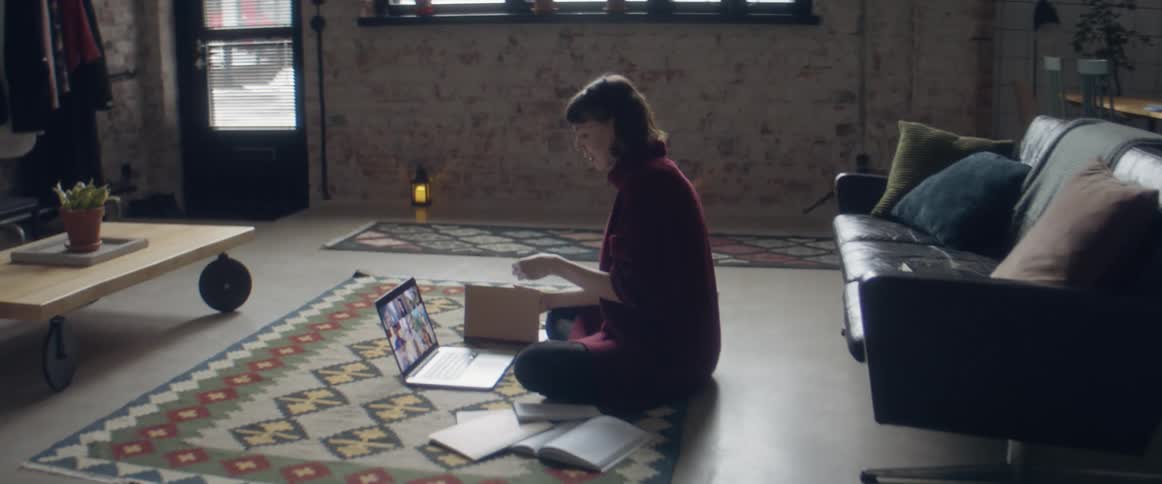 A teenage girl sits on the floor at home and video chats on her laptop