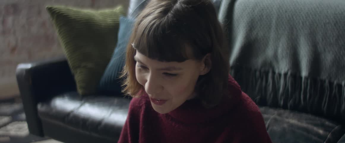A young woman sitting on the floor in front of a couch smiles and waves before talking to someone via video conferencing