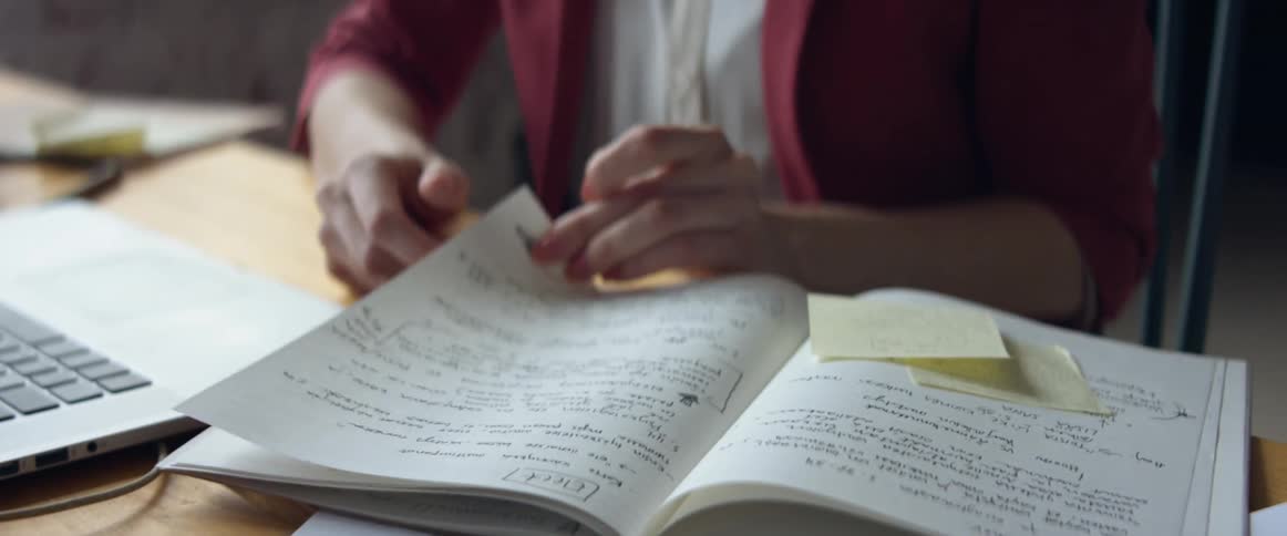 Woman working on a laptop checks her notes on a notebook
