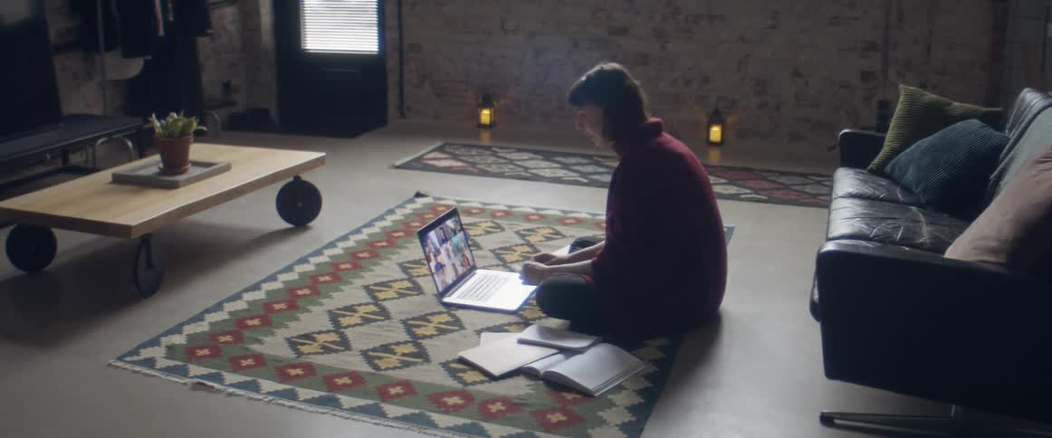 A teenage girl sits on the floor at home and video chats on a laptop