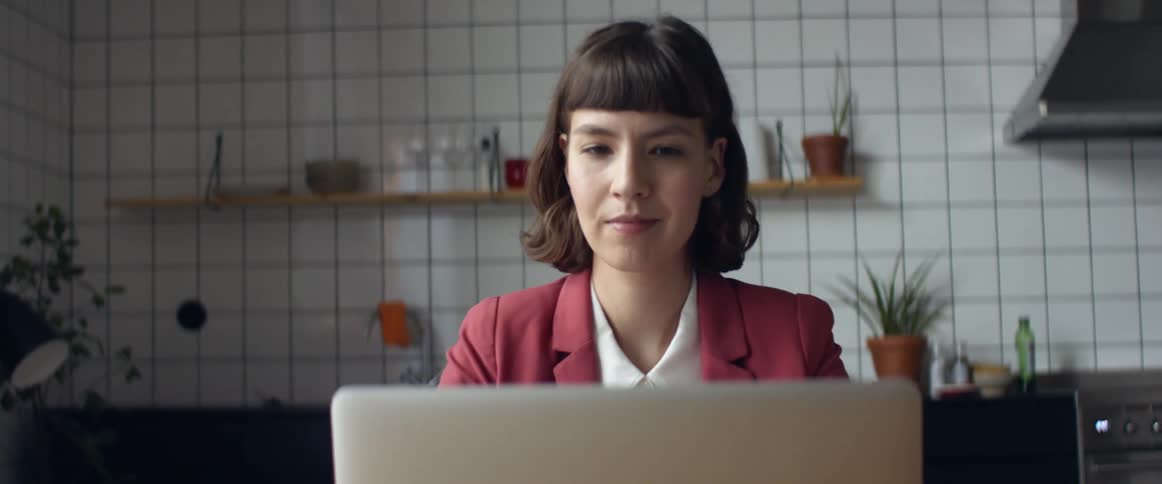 A woman in a suit uses a laptop while sitting at a table in a kitchen during the day
