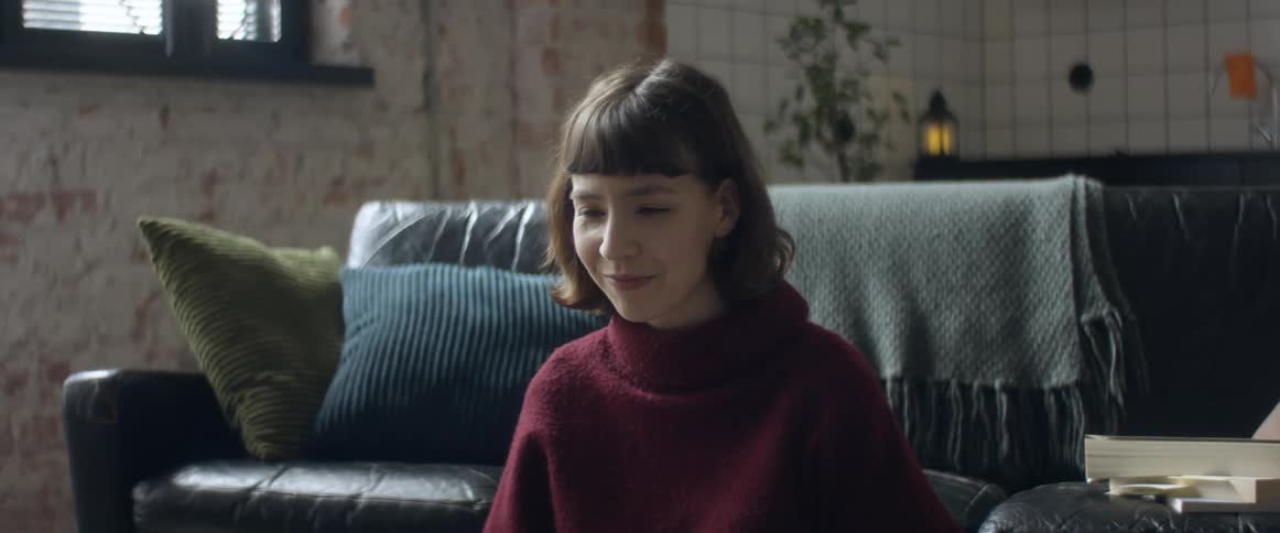 A young woman sitting in front of a couch in a living room talking to someone in front of her