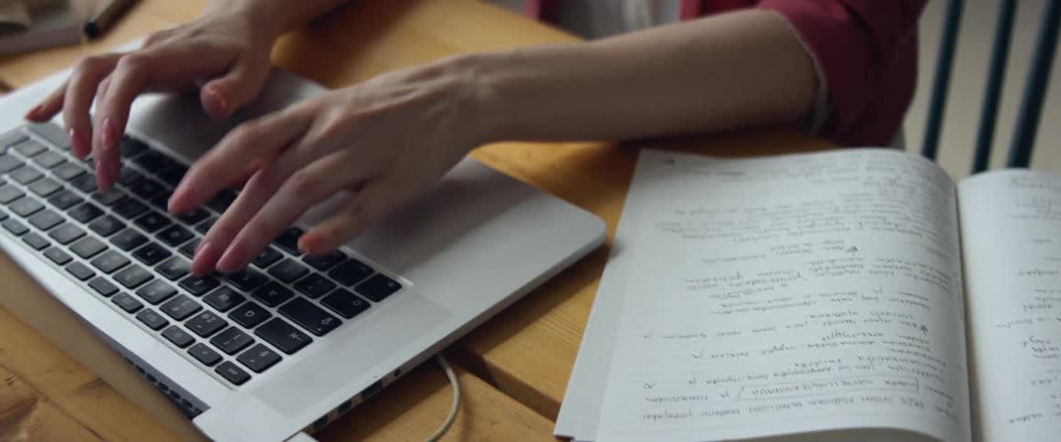 A woman in an office writes in a notebook and then types on a laptop