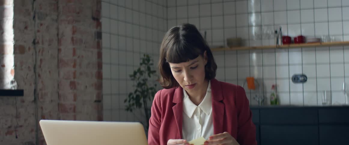 A woman sits at a table in her kitchen with her laptop open in front of her while she looks through notes