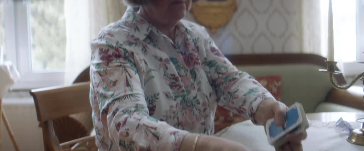 A woman sitting at a table collects scattered playing cards from its surface in a house during the day