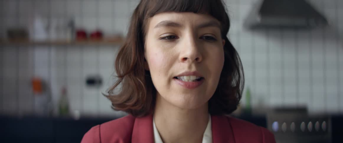 A woman sits in her kitchen and speaks emphatically while gesturing with her hands