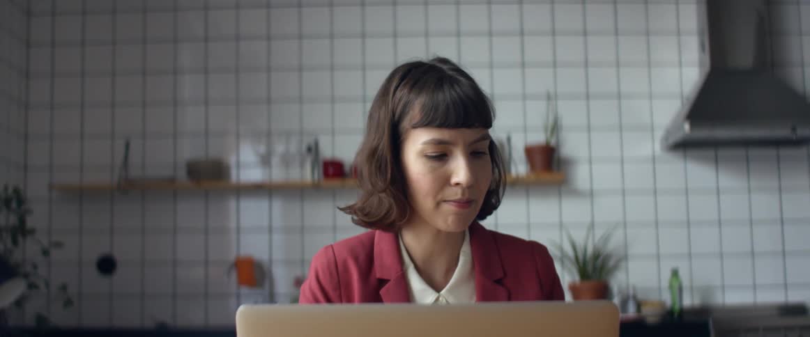 A woman sits in her kitchen while talking and holding up some papers as she participates in a teleconference