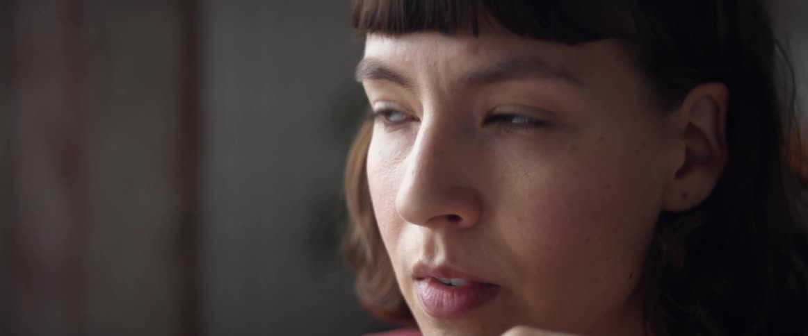 While indoors a woman with brown hair looks into the distance with a contemplative look on her face