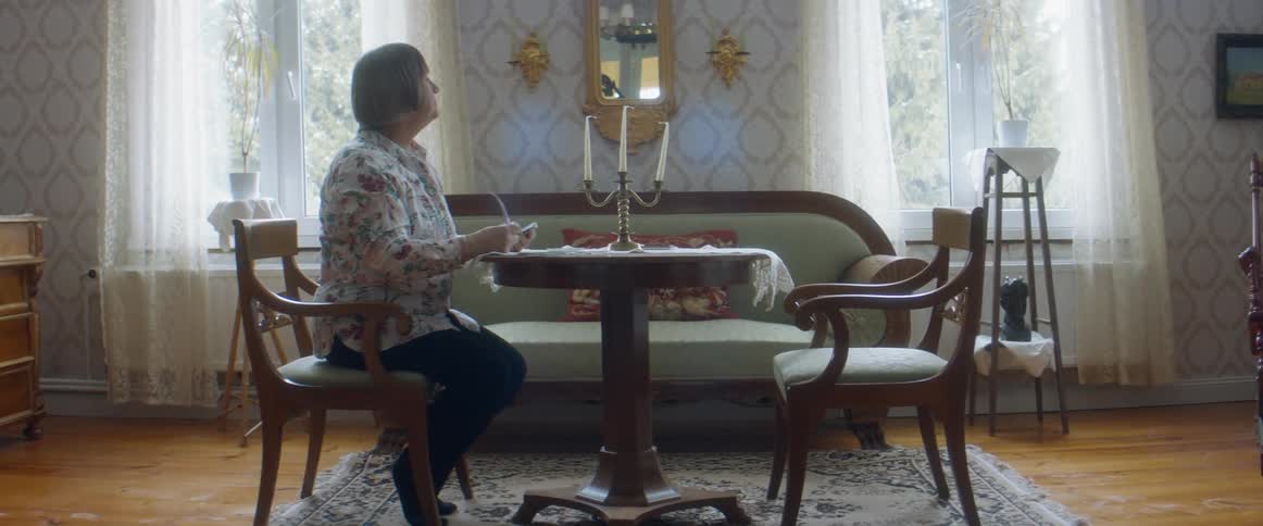An elderly woman sits at a table inside her home and removes her glasses