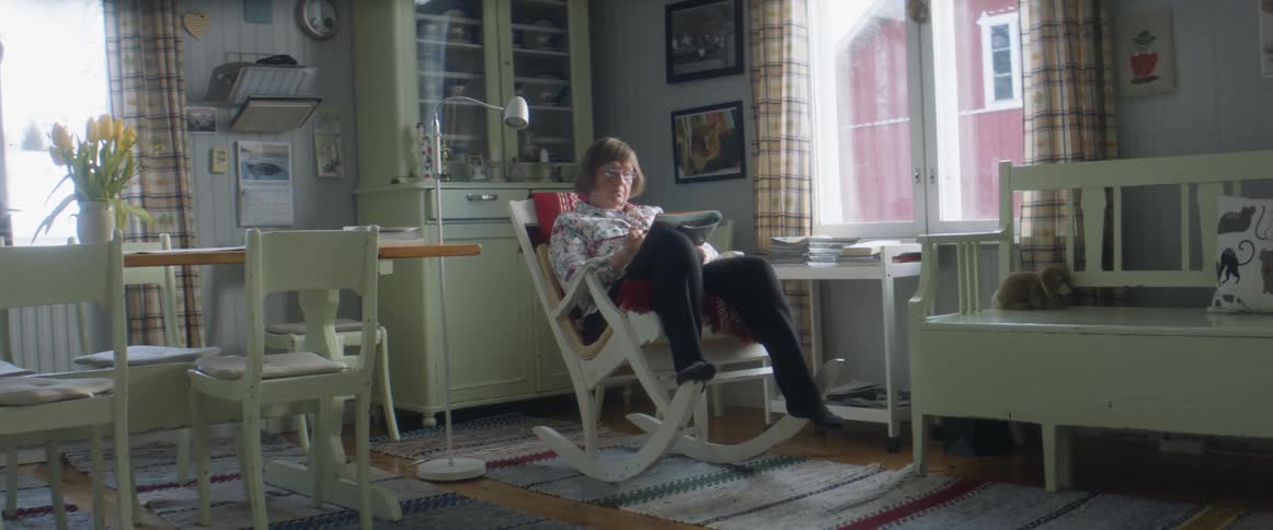 An elderly woman relaxes and sits in a wooden rocking chair while reading