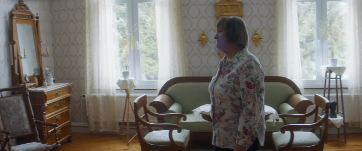 An elderly woman looks out the window in her home and then walks across the room to look at framed photos