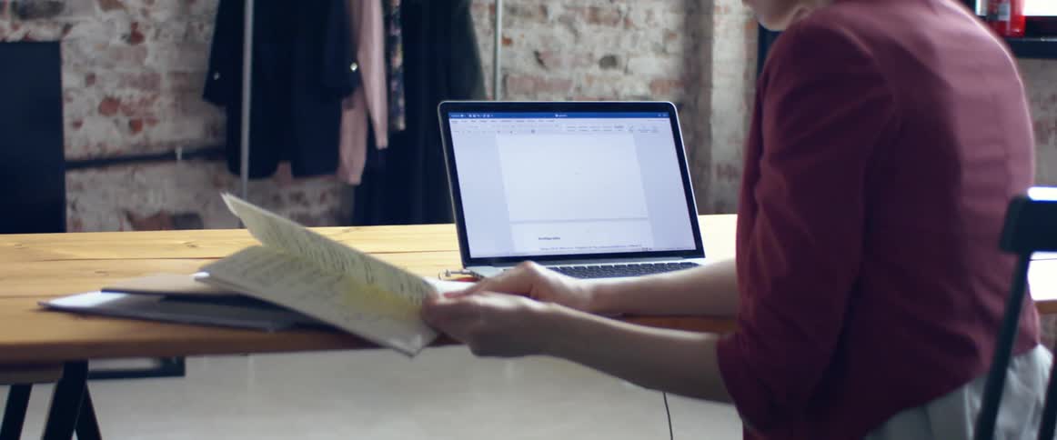 A woman sits at a table with her laptop open as she reviews pages in her notebook
