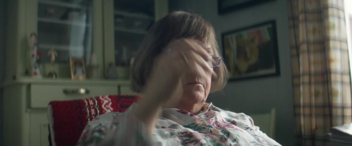 An elderly woman sits at home and takes her glasses off