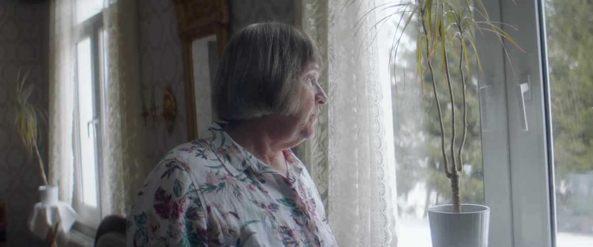 An elderly woman walks through her living room and stops at a window to look outside