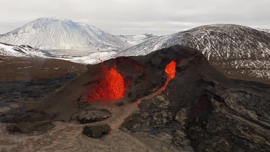 Erupting volcano spews molten hot lava into the air and oozes a river of hot lava with snow covered mountains in the distance