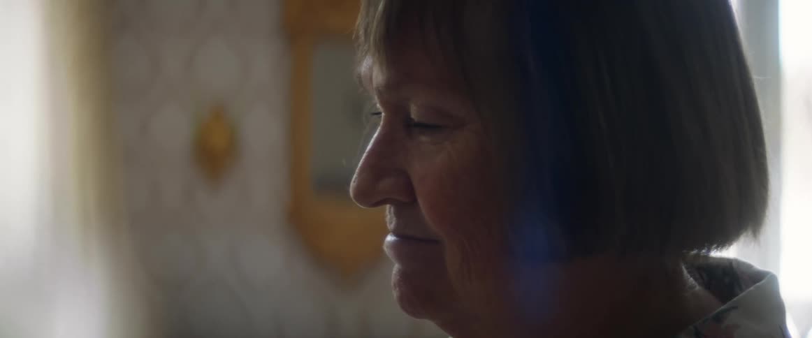 An elderly woman with a short bob smiles as she looks at something in her living room