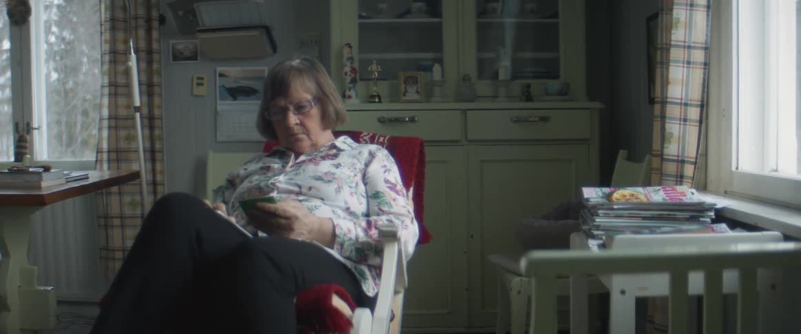 A woman in a room sitting on a rocking chair looking at a smart phone with a wall and window in the background